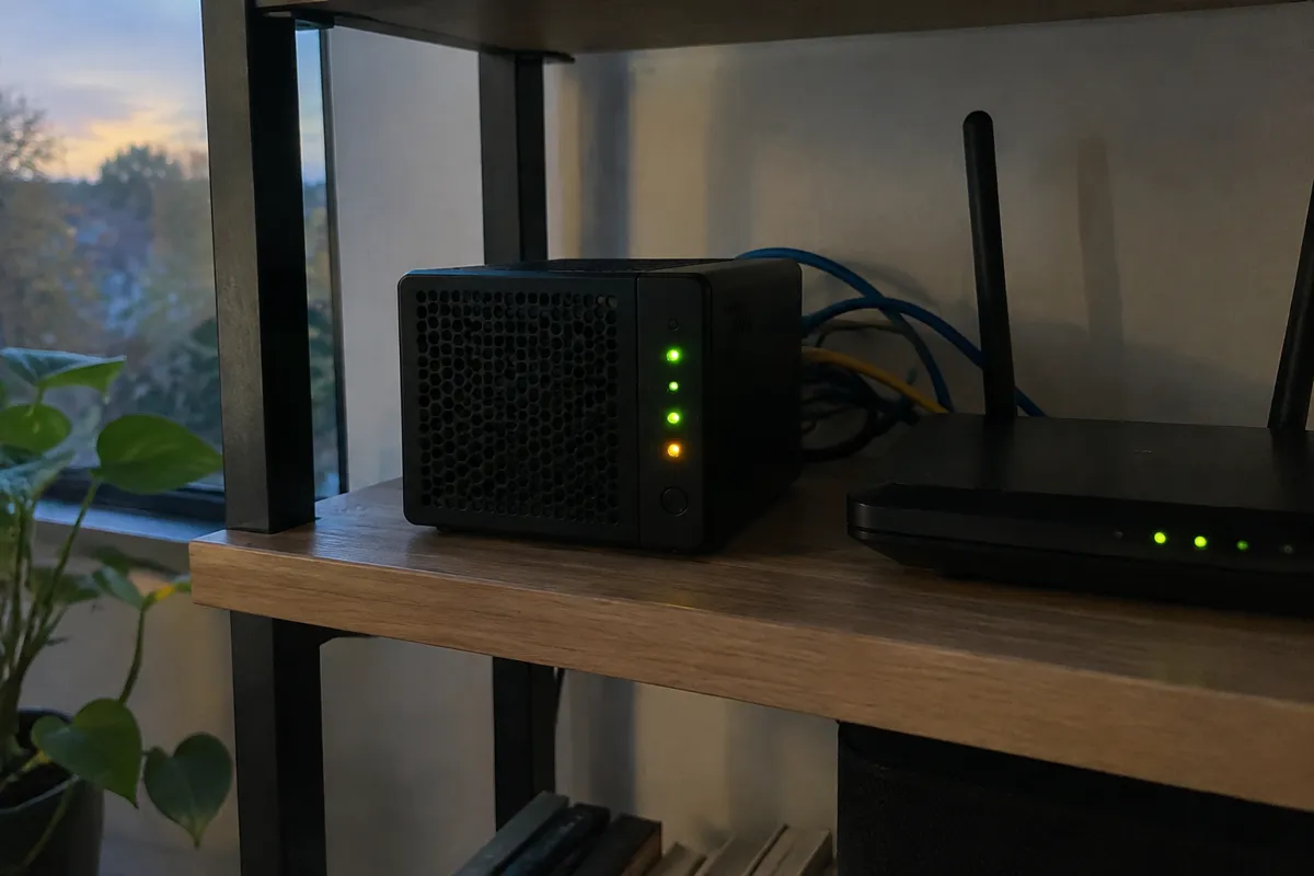 a small black mini server with blinking green and amber LEDs on a shelf, ethernet cables looping behind it, a router beside it, evening ligh