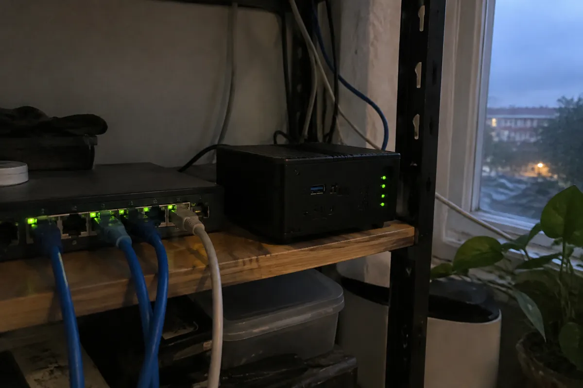 a small mini PC server with blinking green LEDs on a cluttered shelf beside a network switch and a few patch cables, evening light from a ne