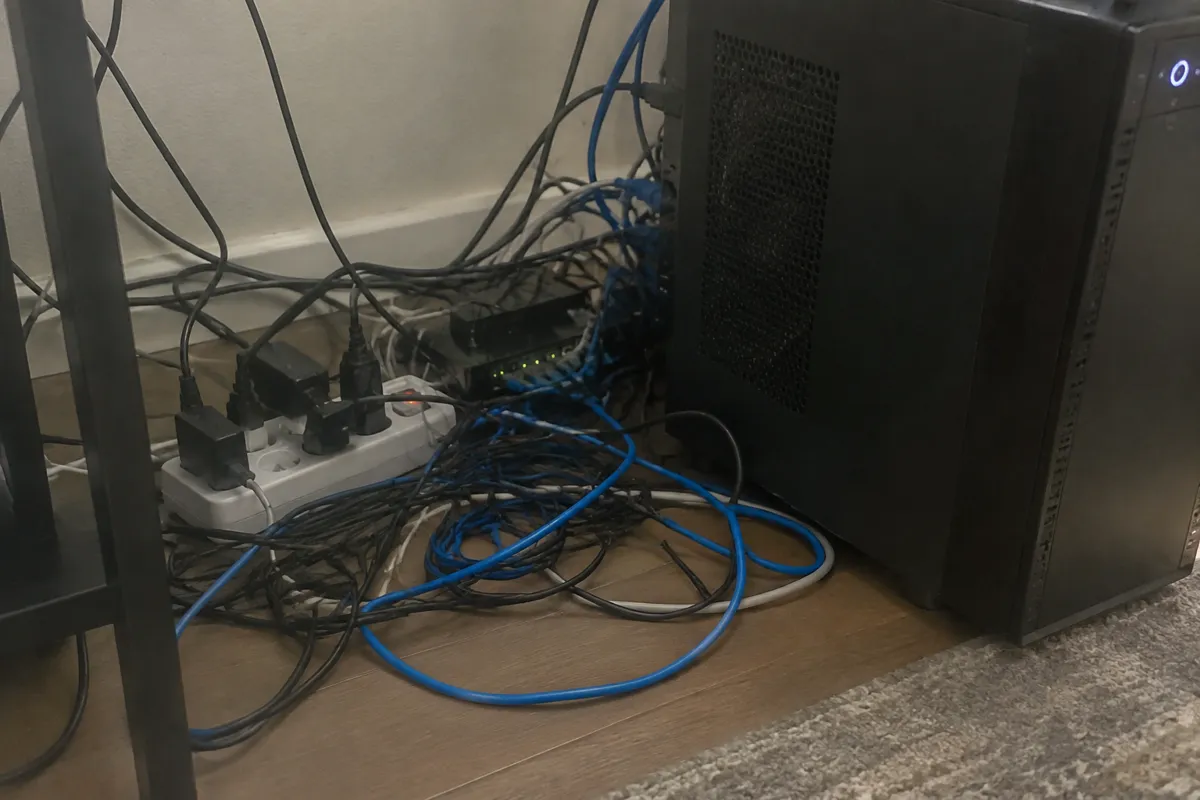 a tangle of ethernet cables and a power strip on a desk floor beside a desktop tower, small LEDs glowing, slightly messy