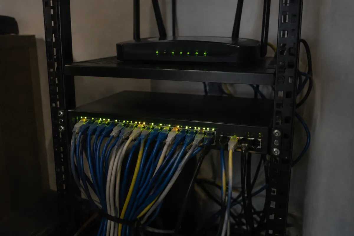 a compact rack-mounted switch with many cables plugged in and glowing port lights, a black router on the shelf above, dim room