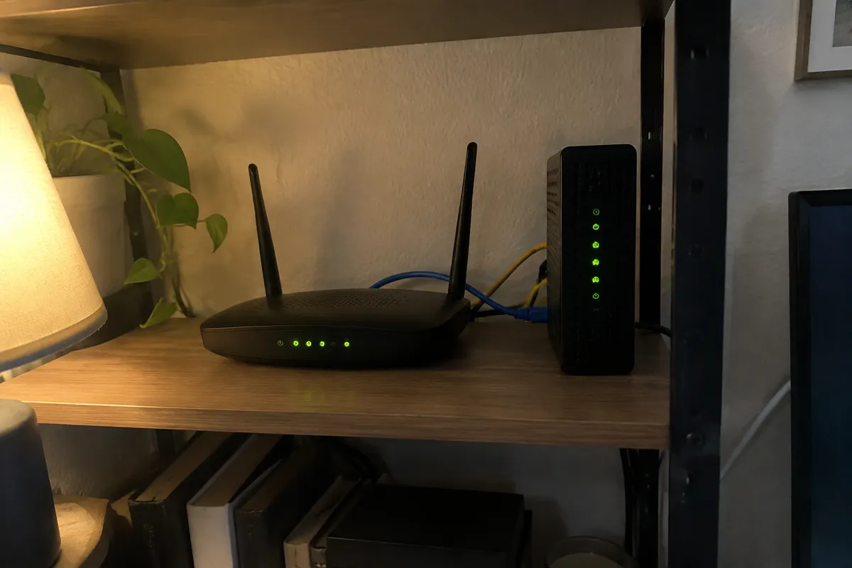 a home router and a modem on a shelf with steady green lights and a couple of ethernet cables, a desk lamp glow nearby