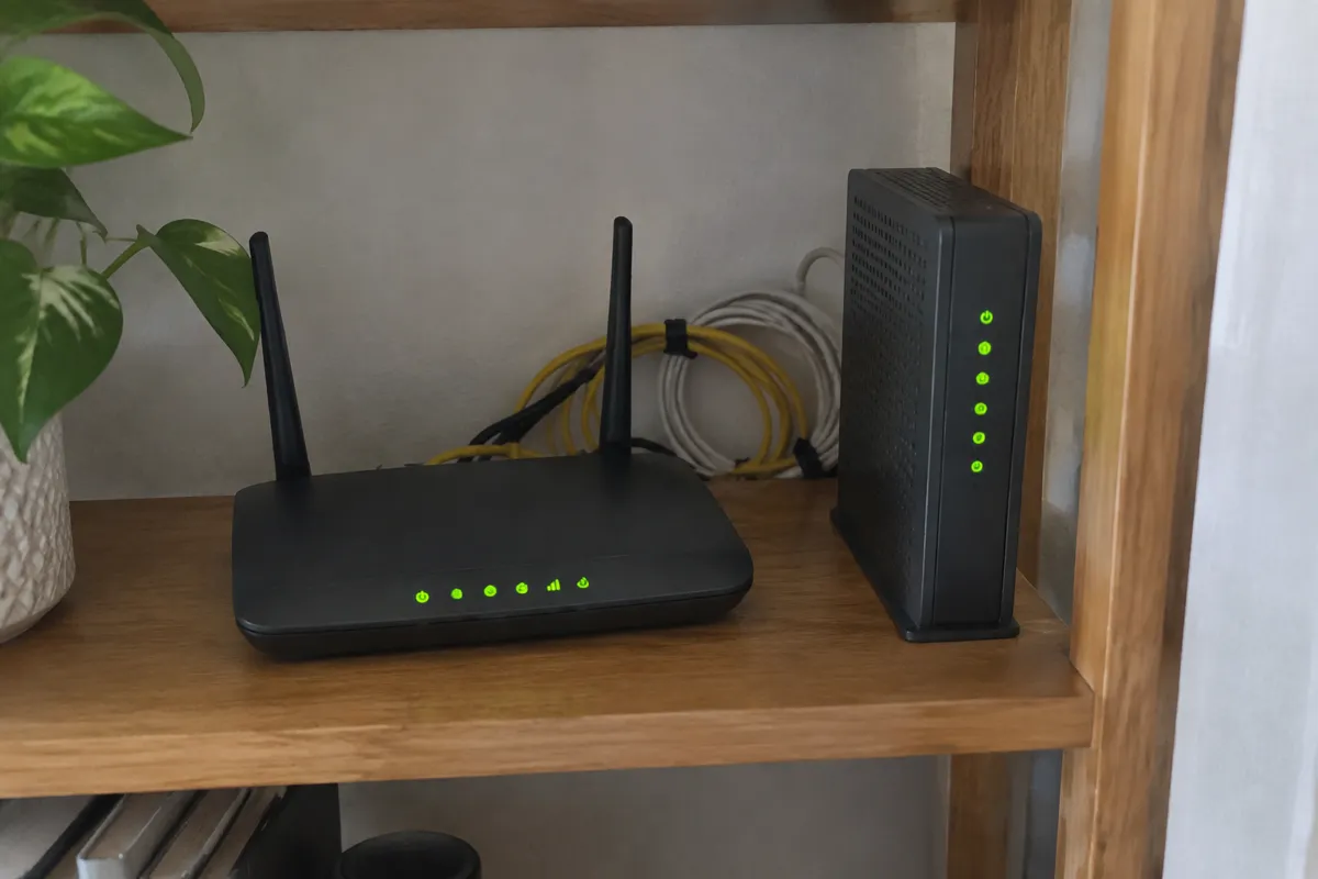a small wifi router and a modem on a wooden shelf with green status lights and neatly coiled cables
