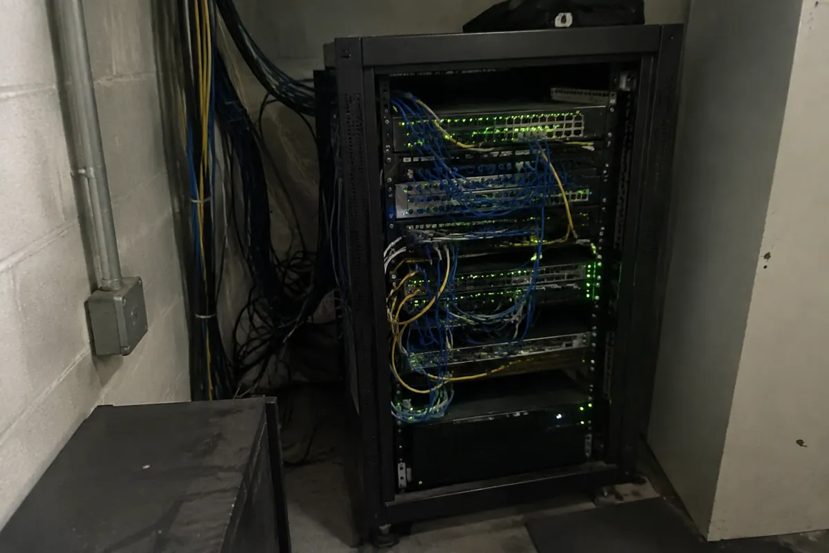 a small server rack with rows of blinking green LEDs and tangled cables behind it, a dim utility room corner