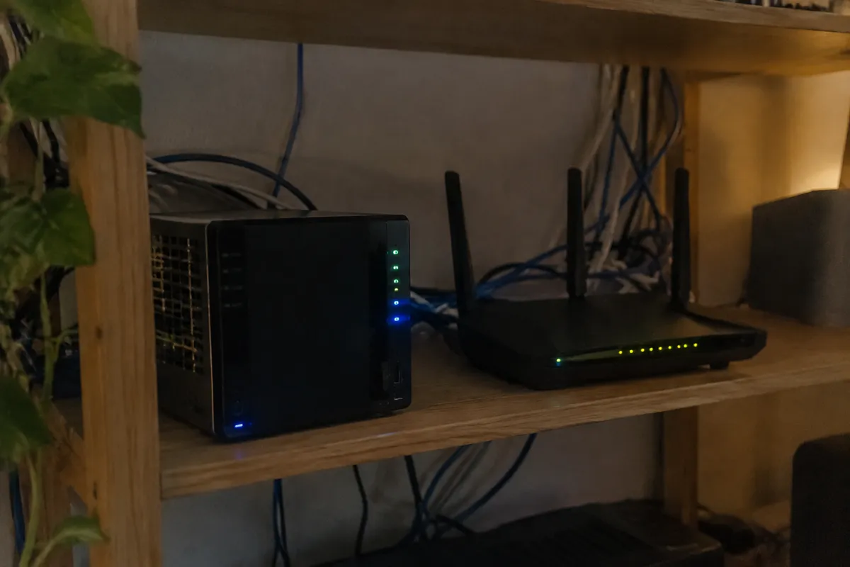a mini home server with blinking LEDs sitting on a shelf beside a router and a tangle of cables, dim evening room