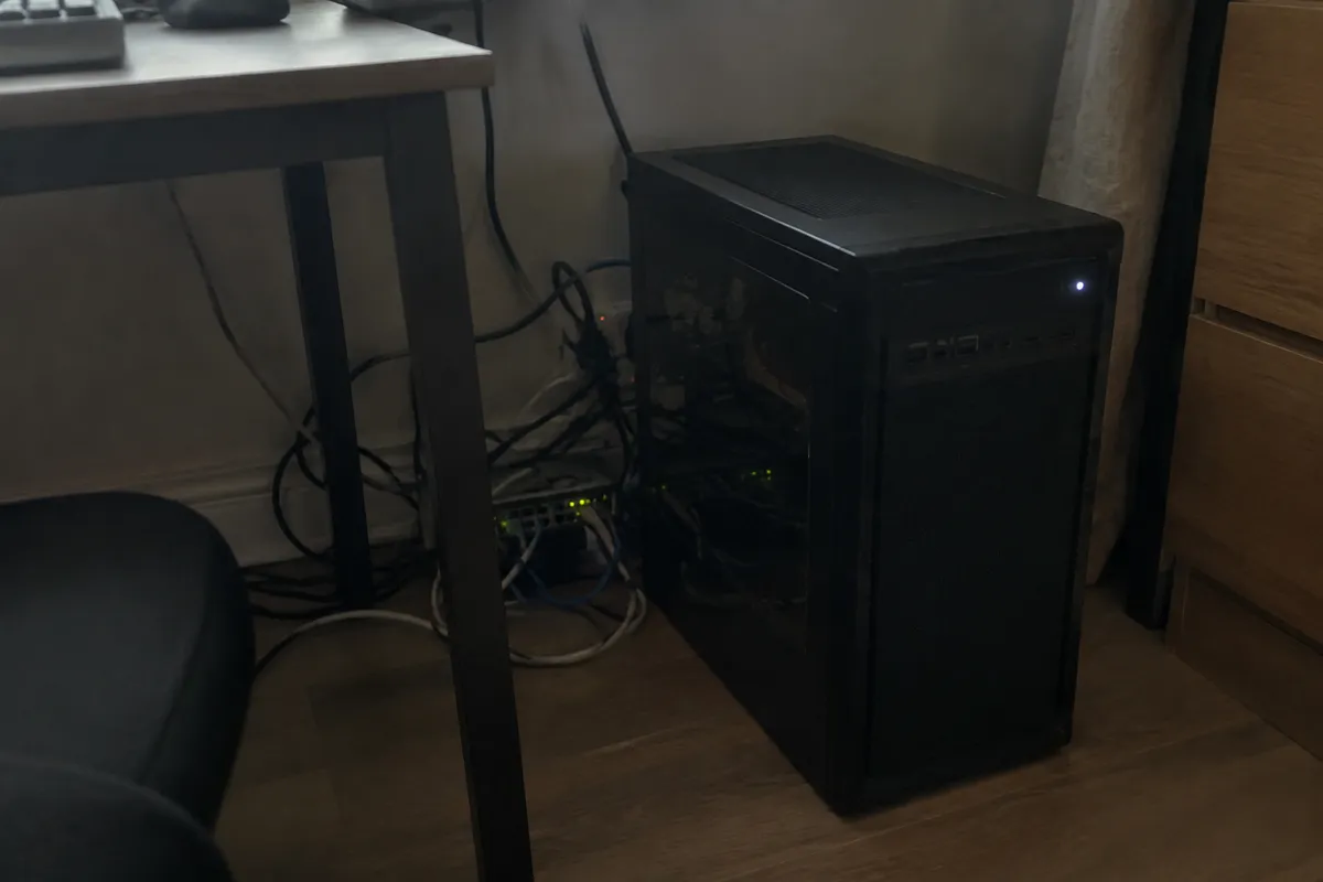 a desktop tower on a floor beside a desk with tangled power and ethernet cables and a small blinking switch, dim room