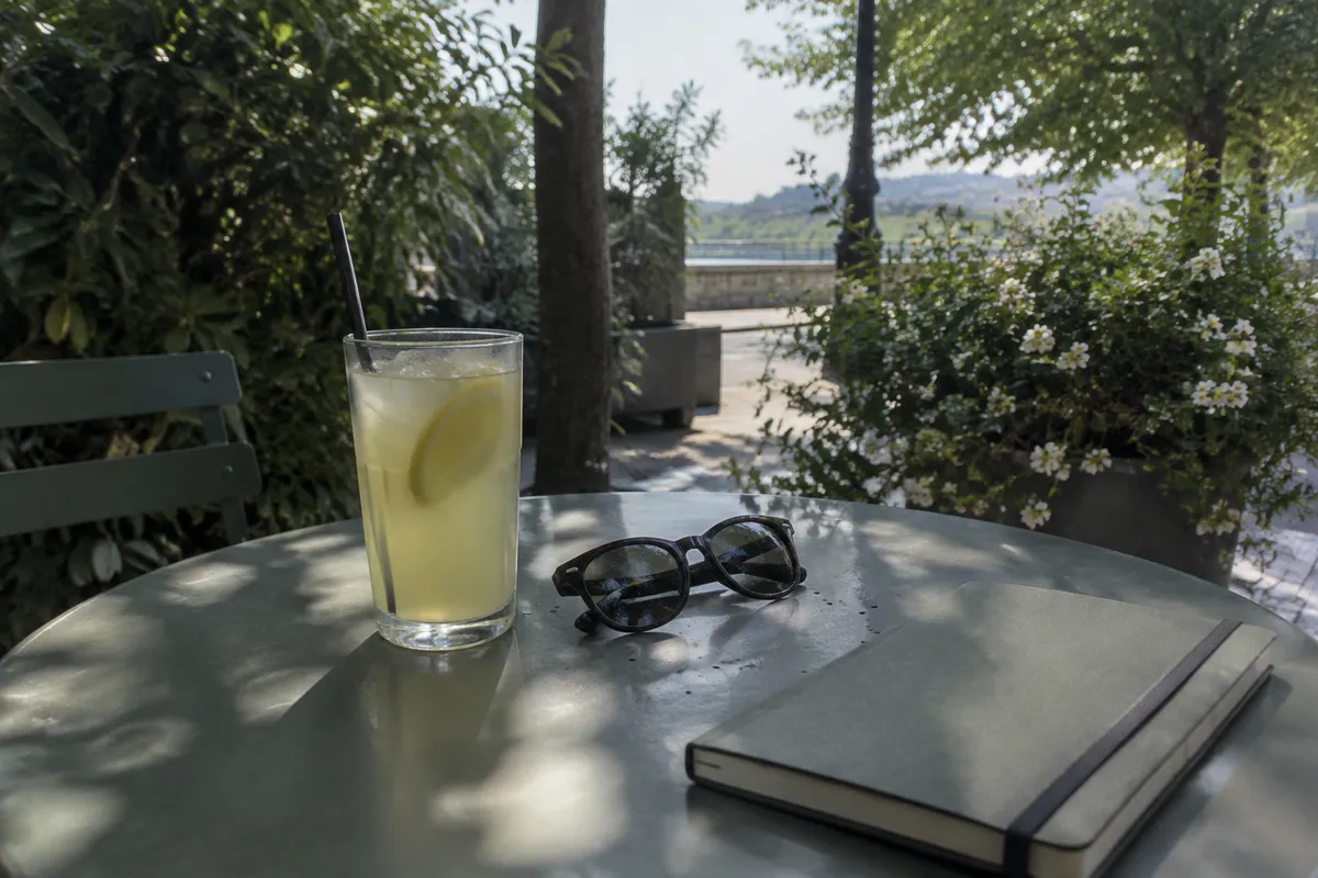 a cafe table outside with a lemonade, sunglasses and a closed notebook in dappled shade