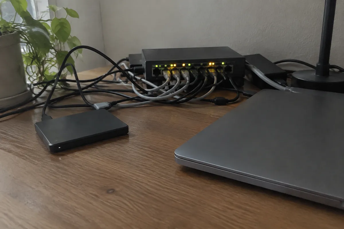 a tangle of cables and a switch with blinking LEDs on a desk beside an external drive and a closed laptop