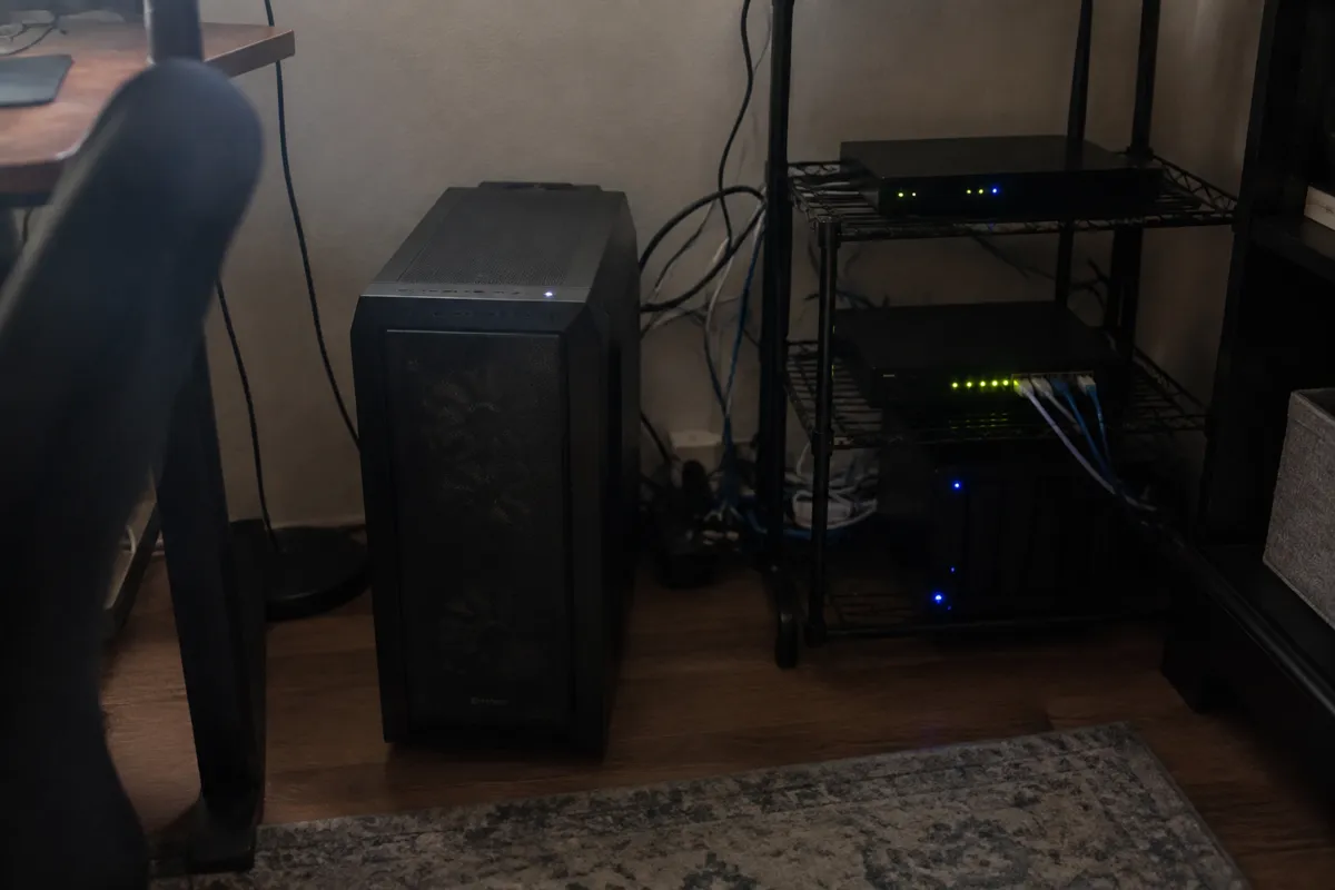 a desktop tower on the floor next to a small rack with blinking LEDs and a tangle of cables, dim home office