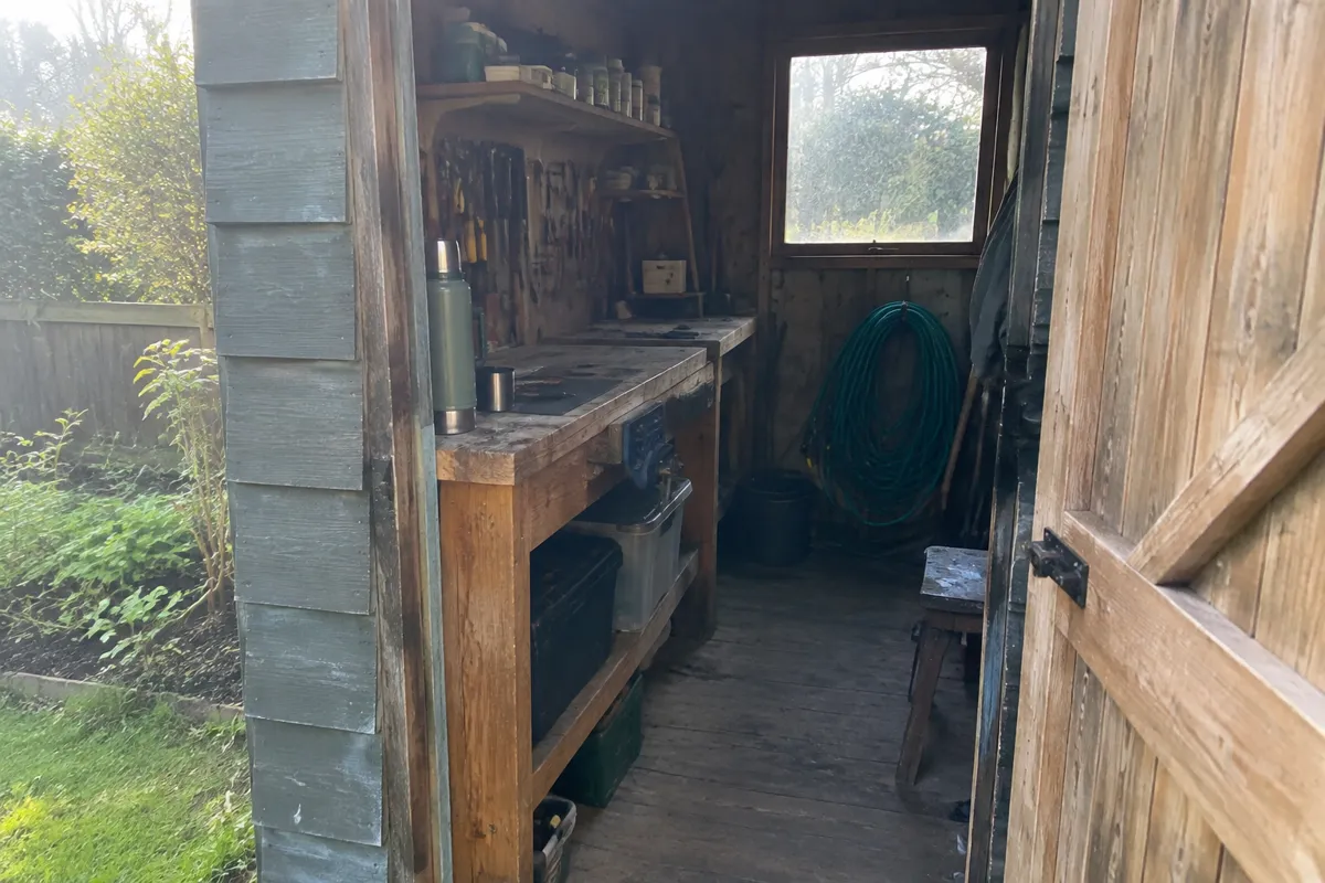 a garden shed doorway with a workbench, a thermos and a coil of garden hose in afternoon light