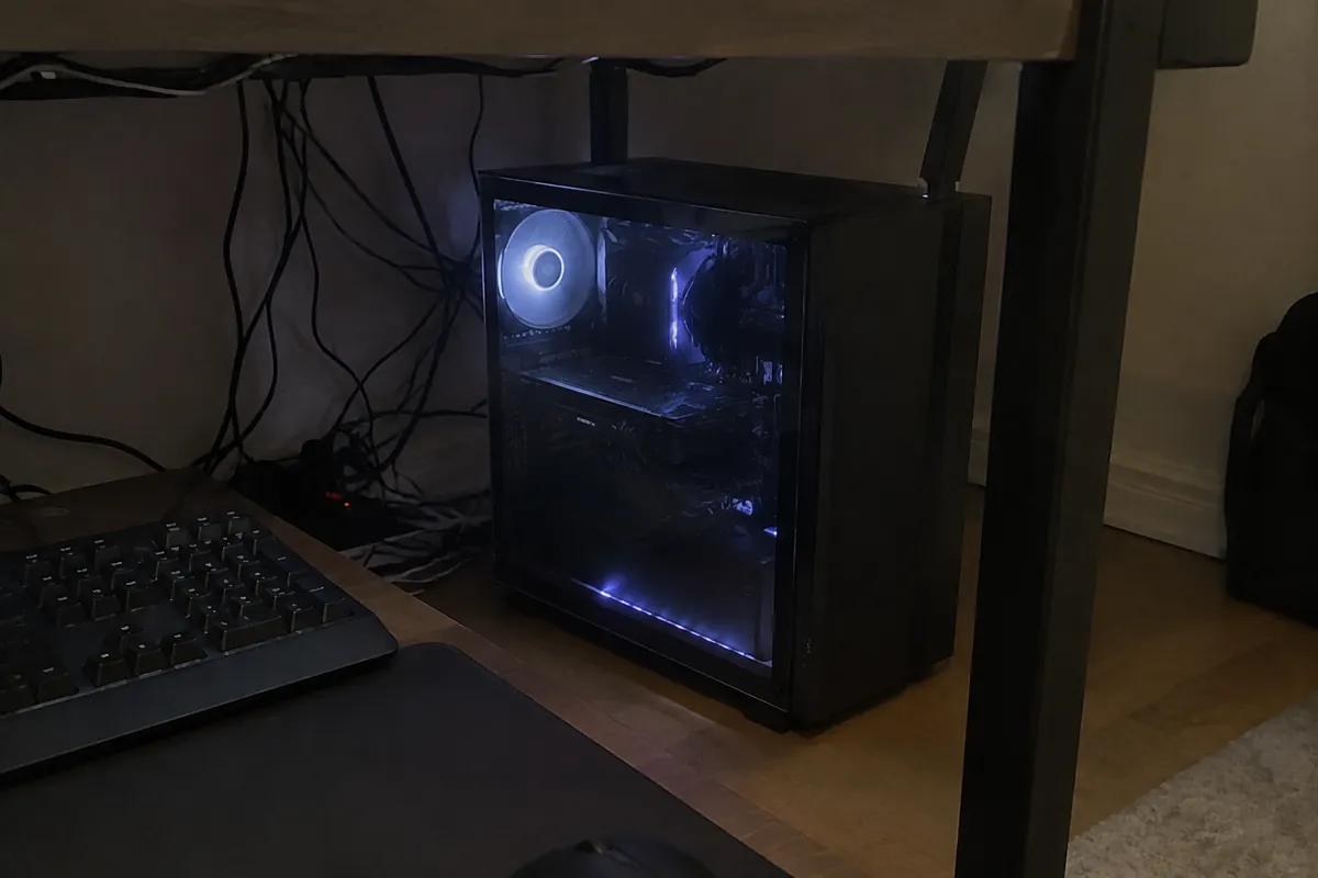 a desktop tower with a glass side panel lit by small LEDs under a desk, a tangle of cables and a keyboard nearby, dim room