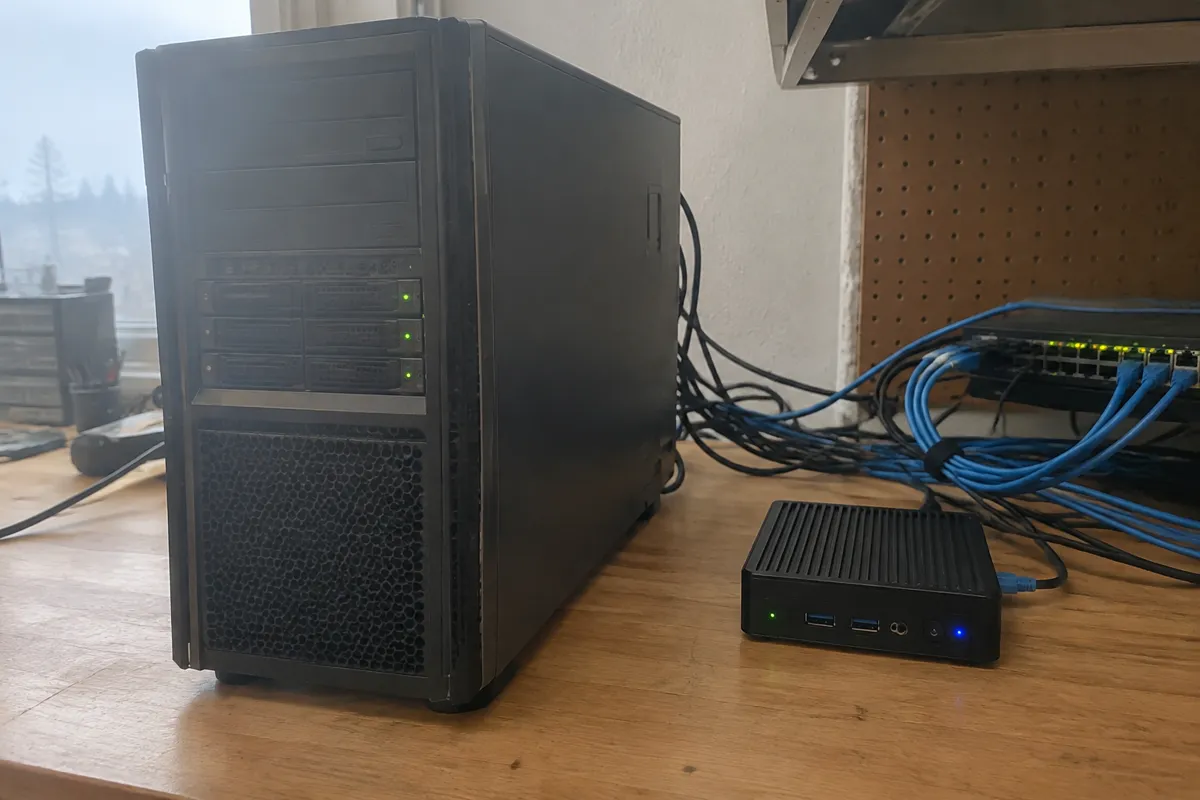 a bulky desktop tower server beside a small mini PC on a workbench with blinking LEDs and cables