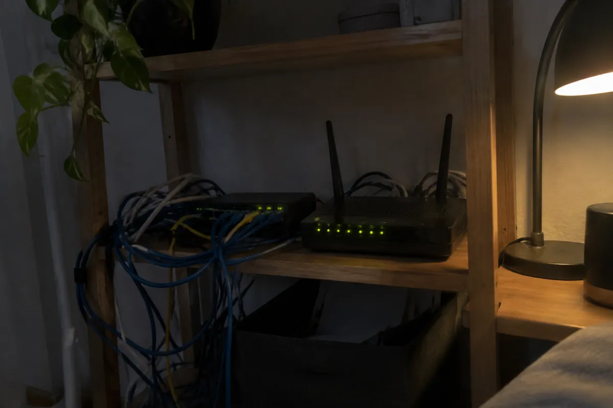 a tangle of ethernet cables and a router with glowing lights on a shelf, a small desk lamp nearby, dim room