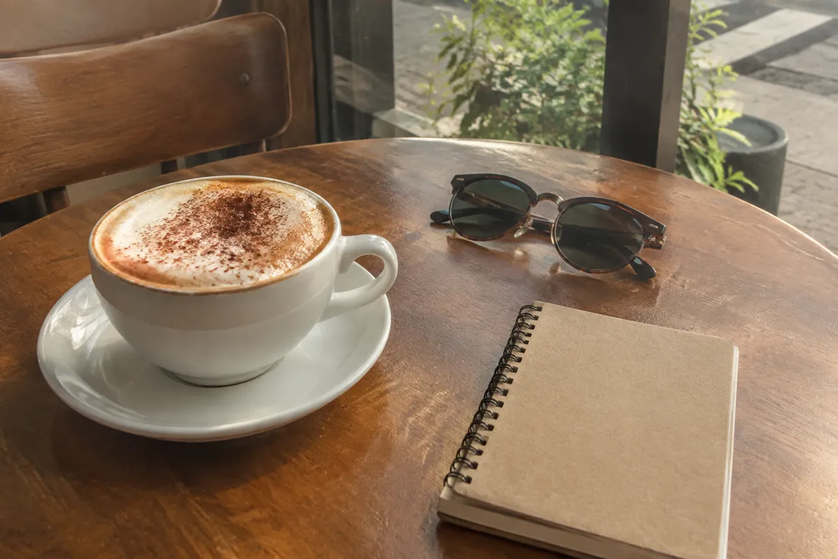 a cafe table with a cappuccino, a small notebook and a pair of sunglasses in warm light
