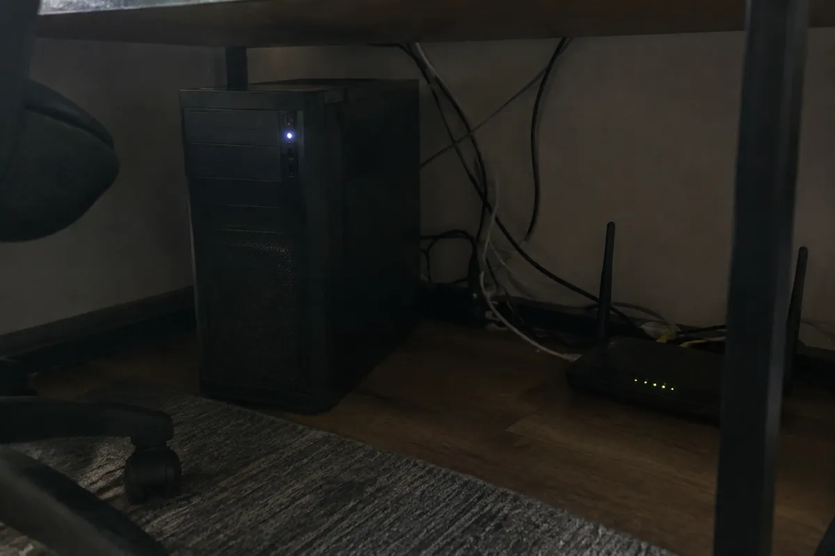 a desktop tower humming under a desk with a small status light and a few cables running to a router, dim room