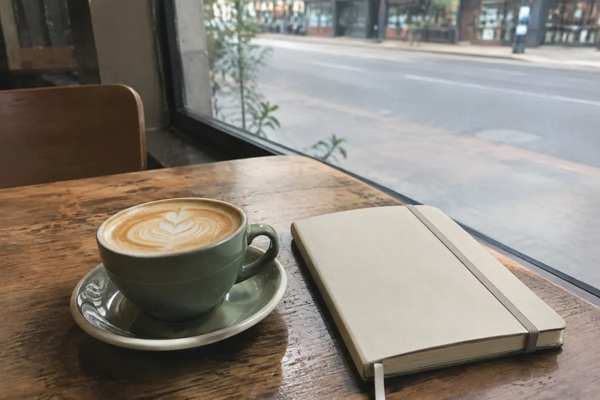 a cafe table with a flat white and a notebook, soft window light, blurred street outside