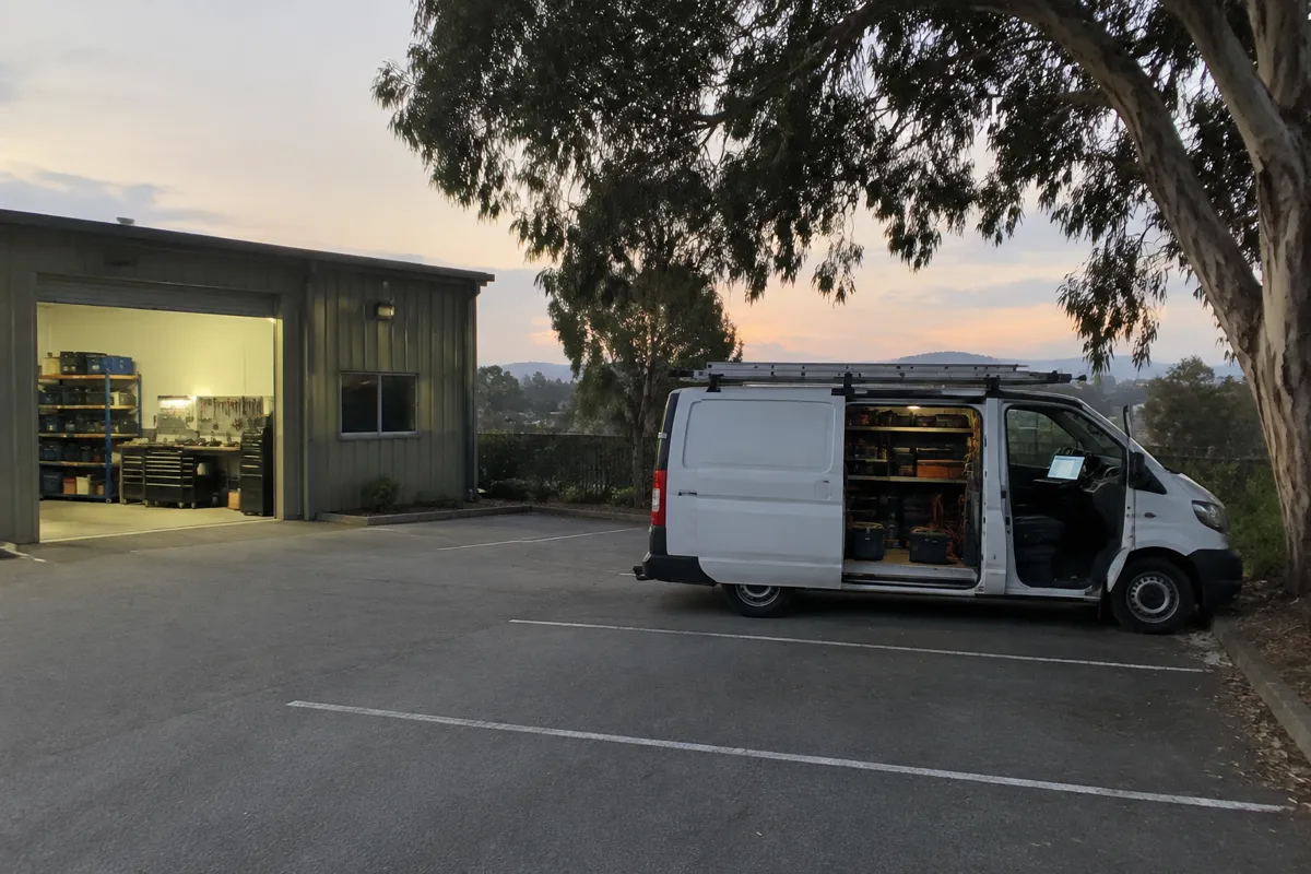 an electrician's van parked outside a small workshop at dusk with a laptop glowing on the dashboard