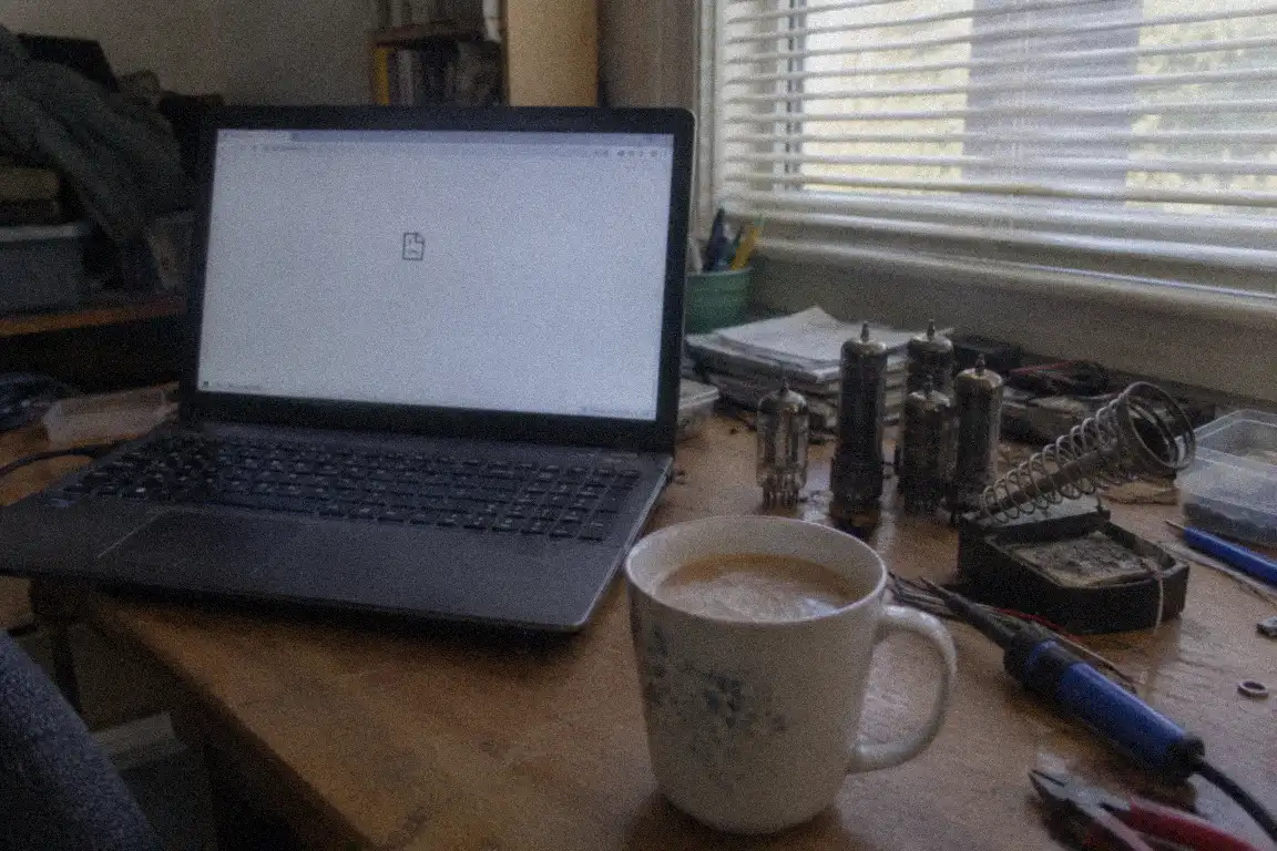 a laptop screen glowing in a dim spare room, with a mug and a pile of papers on the desk