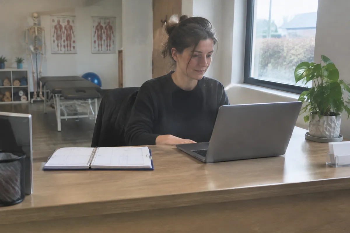 a physiotherapist at her clinic reception desk reading an email on a laptop