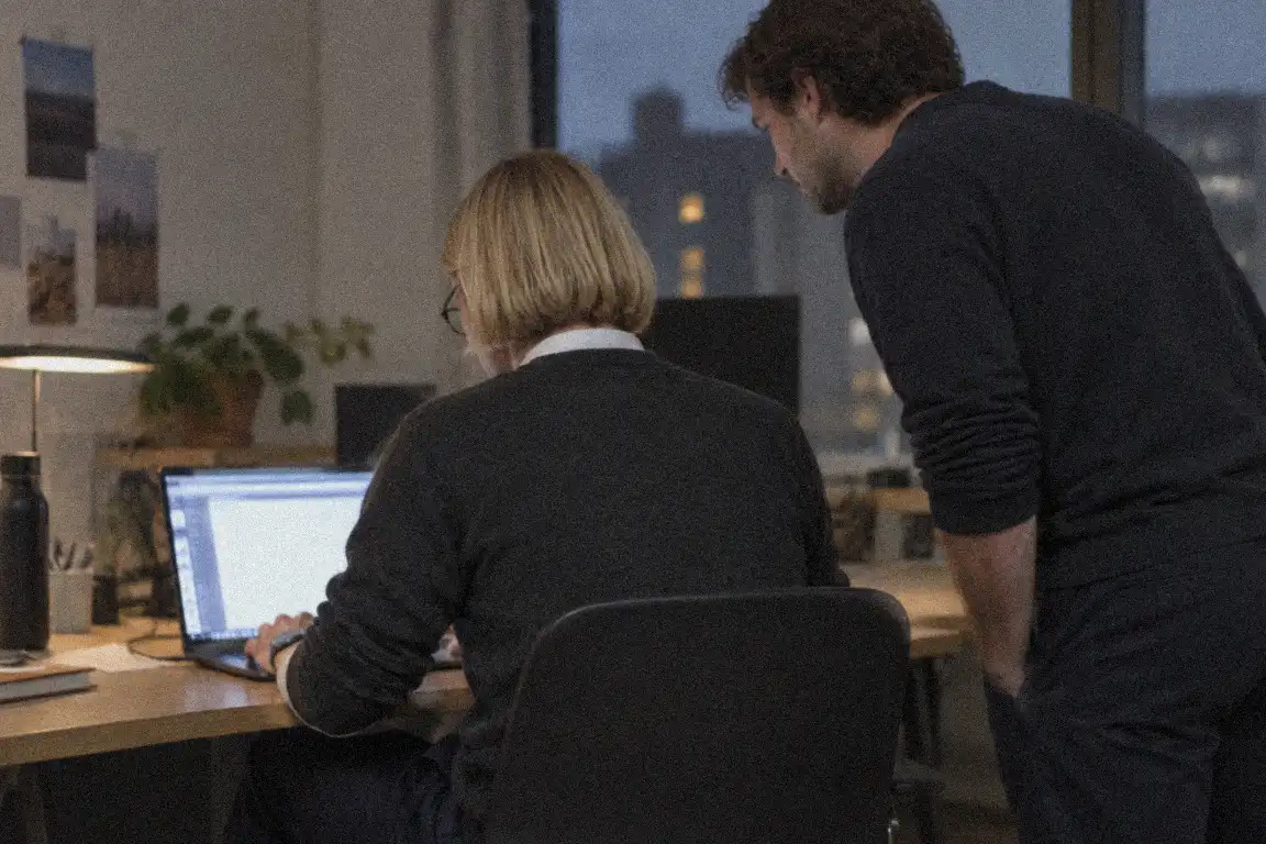 two people at a shared desk looking at a laptop screen in a dim office