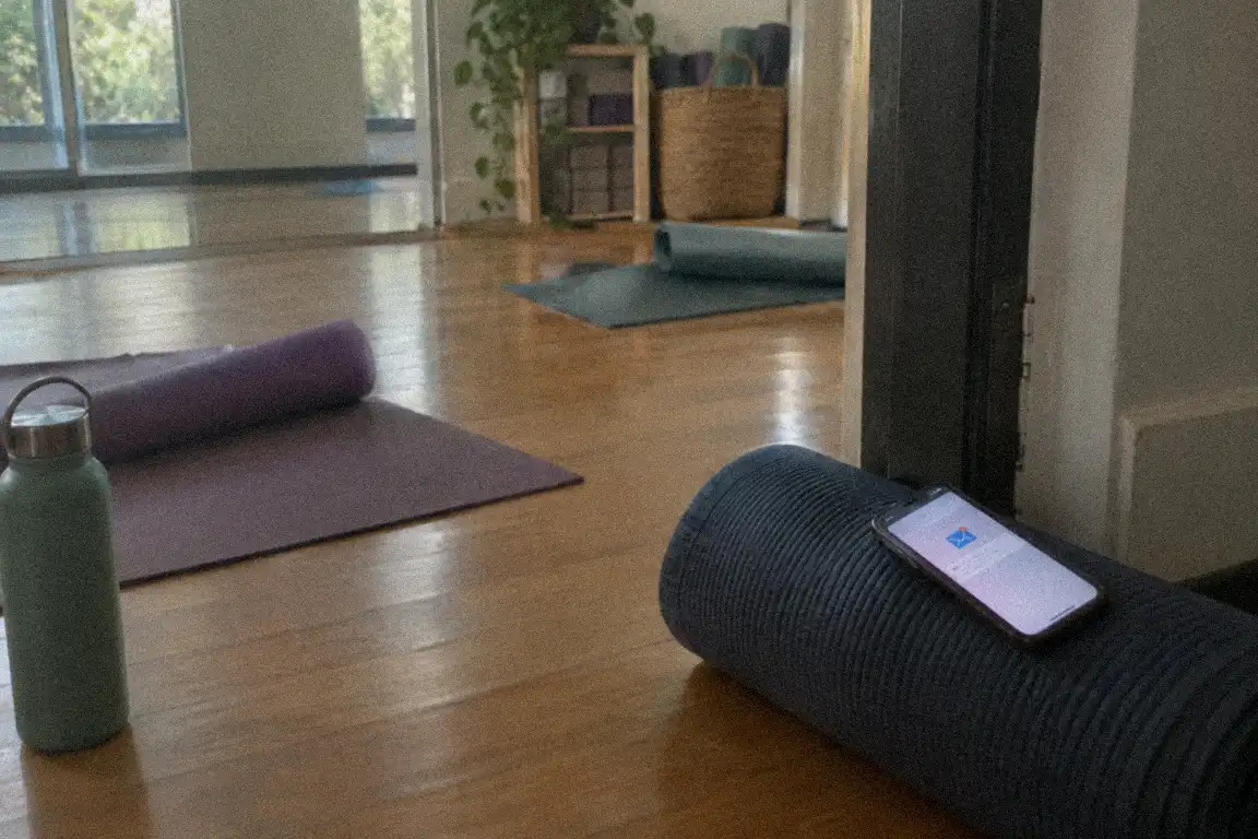 a woman in a quiet yoga studio reading her phone, mats stacked behind her in soft daylight